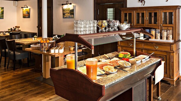 Un buffet de petit-déjeuner proposant une variété d’aliments et de boissons, un buffet en bois doté de portes en verre, et un espace repas avec des chaises et des tables.