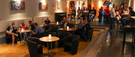 A restaurant with round tables and black chairs, featuring a mix of seated and standing patrons.