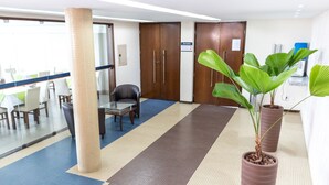 Un vestíbulo de hotel con un estampado de cuadros en el suelo, una pared de cristal, una zona de estar con mesas y sillas, y dos plantas grandes en macetas.
