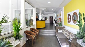 Un área de recepción con pared amarilla, mostrador de recepción, sillas de mimbre, un sofá y plantas en macetas.