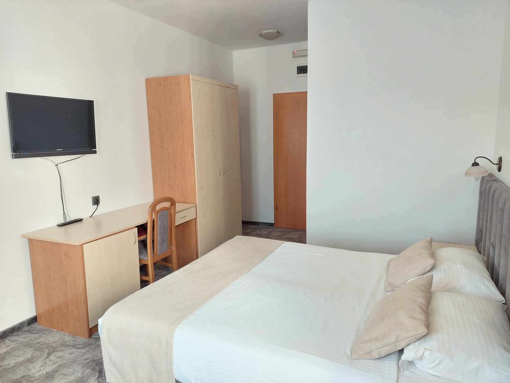 Habitación de hotel con cama, escritorio, silla, armario, televisor y lámpara de pared.