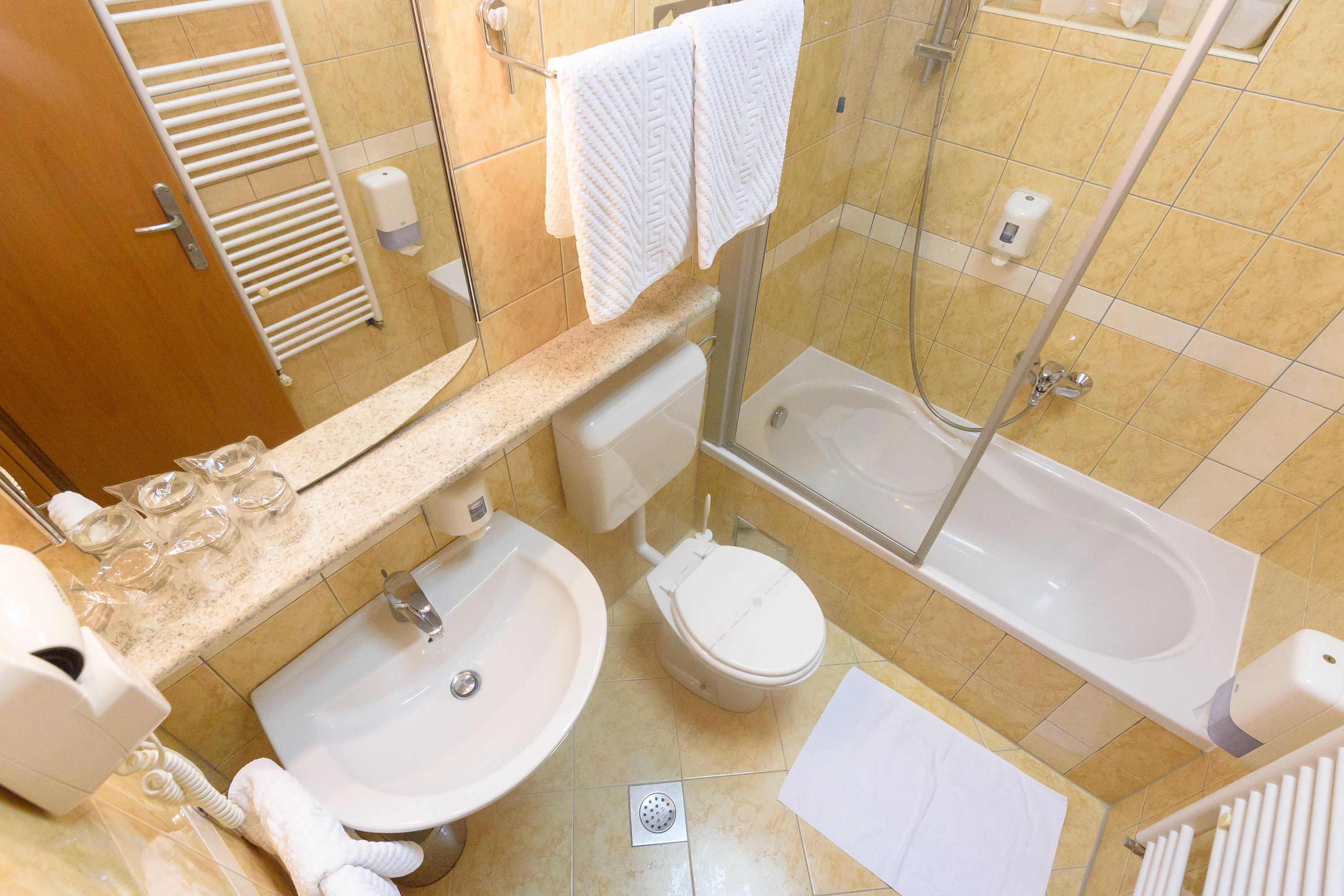 Baño con lavamanos, inodoro y bañera. Toallas colgadas en un perchero, dispensador de jabón y cabina de ducha con mampara de vidrio.