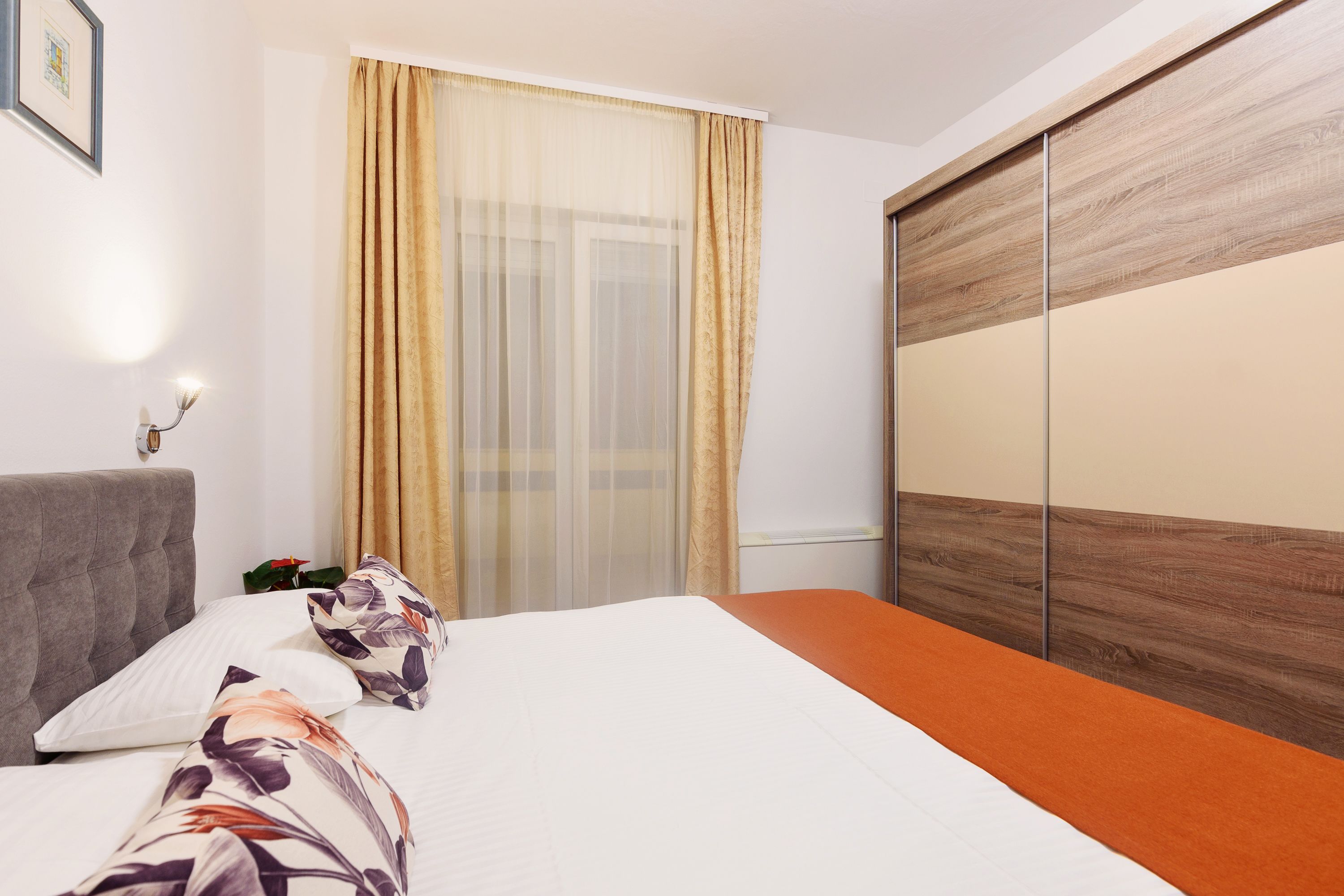 A hotel room with a bed, a grey headboard, a wooden wardrobe, and a window with sheer curtains.