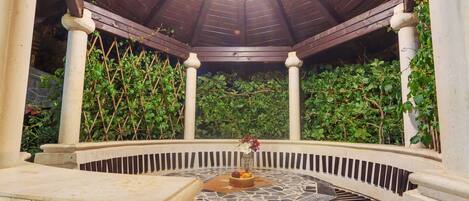 A gazebo with a tiled floor, surrounded by greenery and stone walls.