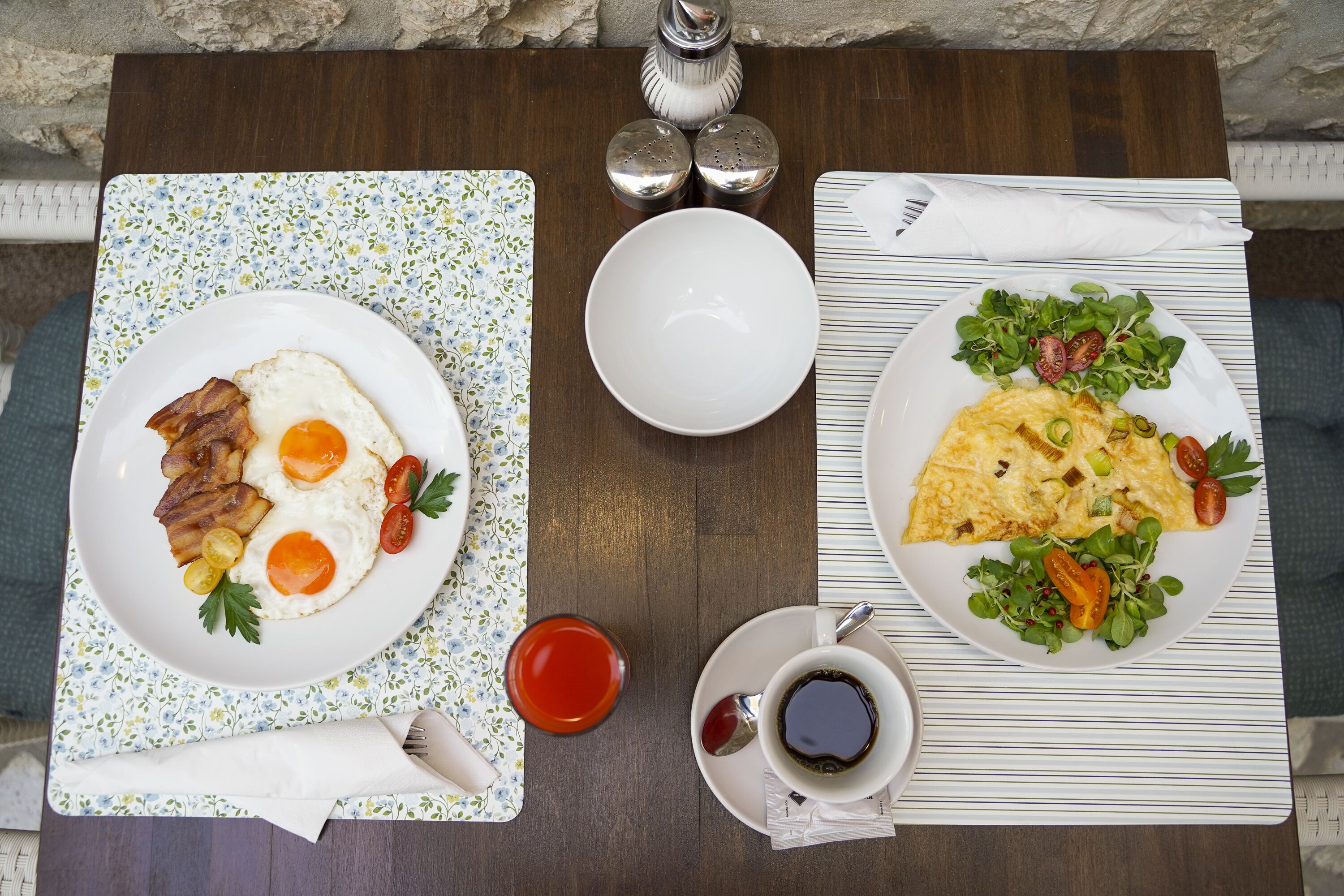 Une table de petit-déjeuner avec des assiettes d’œufs, de bacon et de légumes verts, une tasse de café, ainsi que des salières et des poivrières.