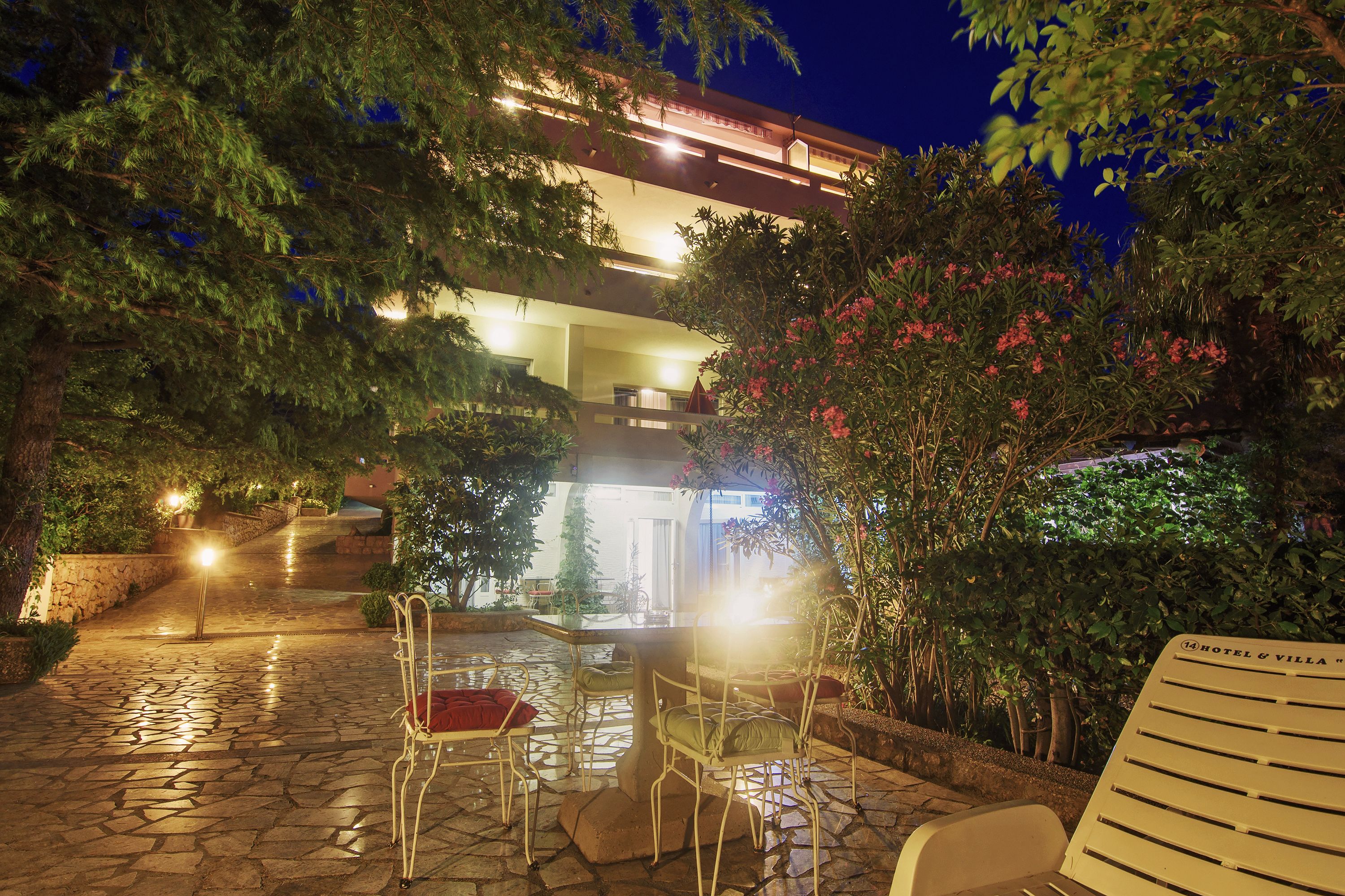 Un patio de hotel por la noche con suelo de piedra, mobiliario para exteriores y abundante vegetación.