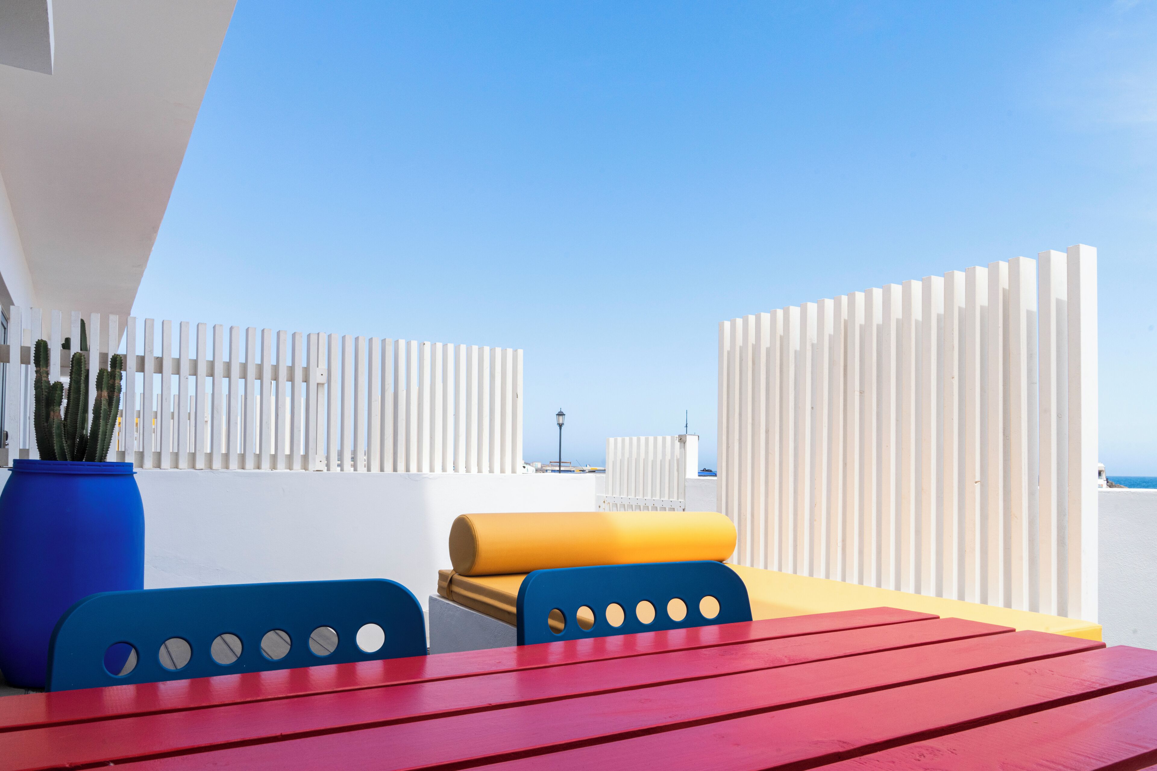A red table with blue chairs, a yellow cushion, and a blue vase on a balcony with white railings and a clear sky.