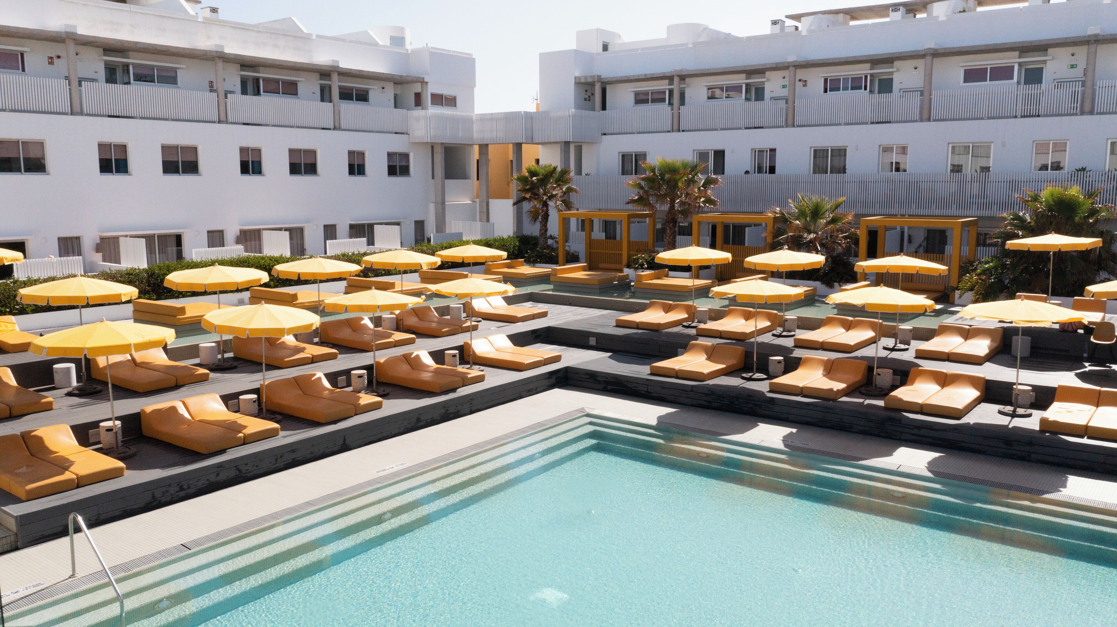 Un espace piscine aménagé sur le toit, doté de plusieurs chaises longues et de parasols, et entouré d’immeubles blancs.