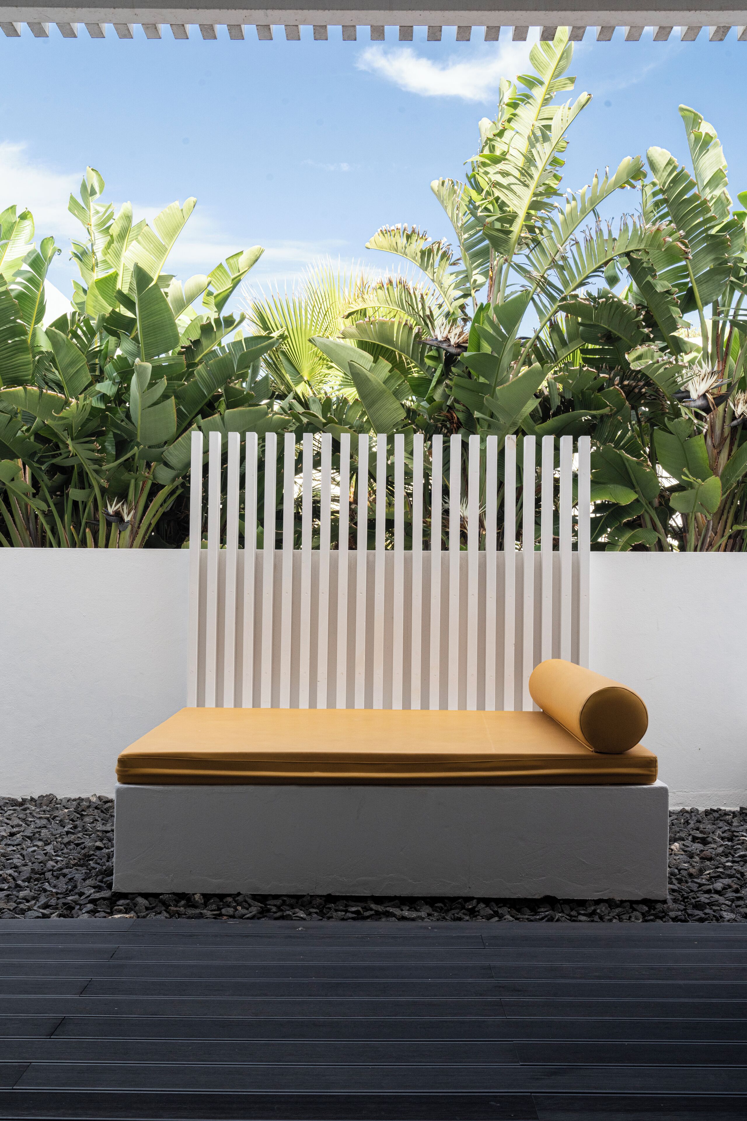 A modern outdoor seating area with a white slatted bench and a yellow cushion, surrounded by lush tropical plants.