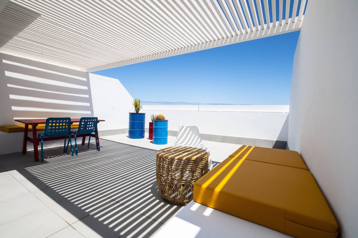 Un espace extérieur moderne aménagé avec une table et des chaises, un pouf en osier et des plantes en pot.