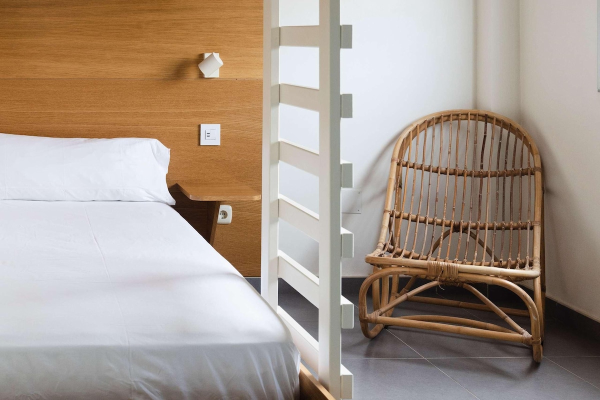 A bunk bed with a wooden headboard, a white mattress, and a white side rail. There is a wicker chair and a woven basket hanging from the ceiling.