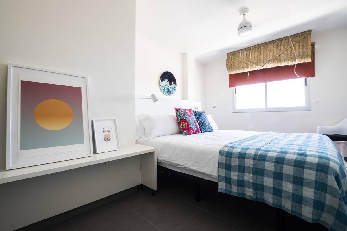 Une chambre à coucher comprenant un lit, une table de chevet, un tableau encadré, une horloge murale et une fenêtre agrémentée d’une lambrequin.