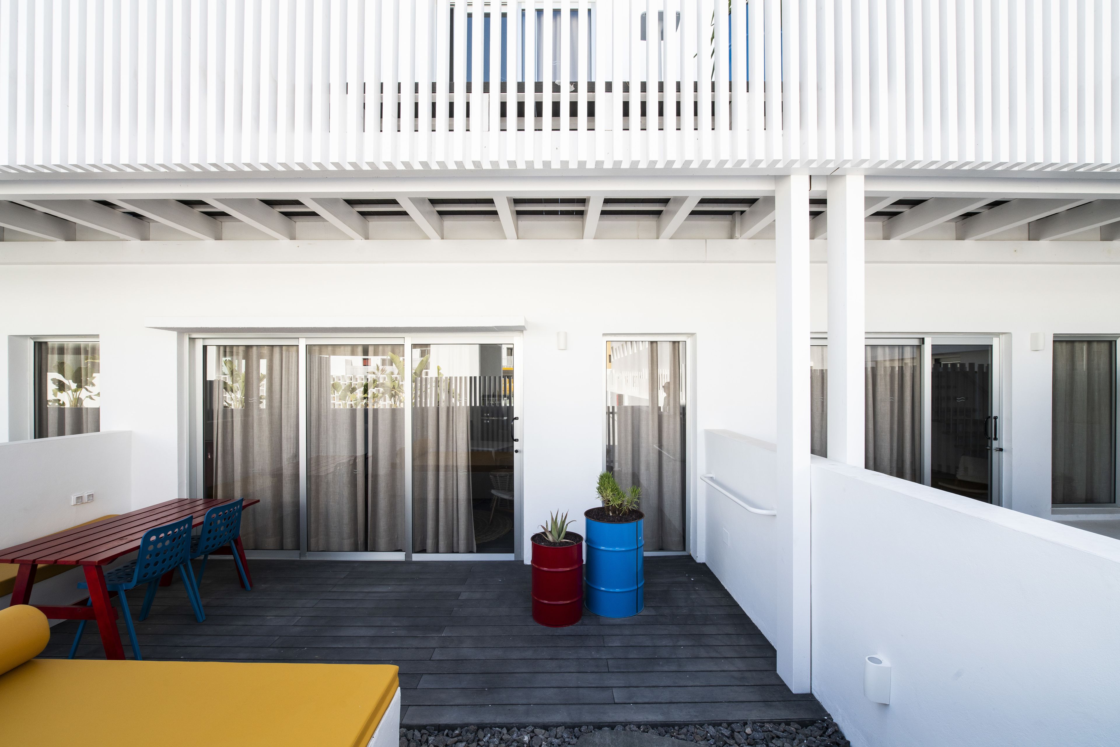 A covered patio with a wooden table and chairs, a red and blue barrel planter, and a white wall.