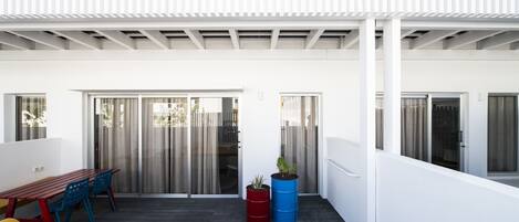 A covered patio with a wooden table and chairs, a red and blue barrel planter, and a white wall.