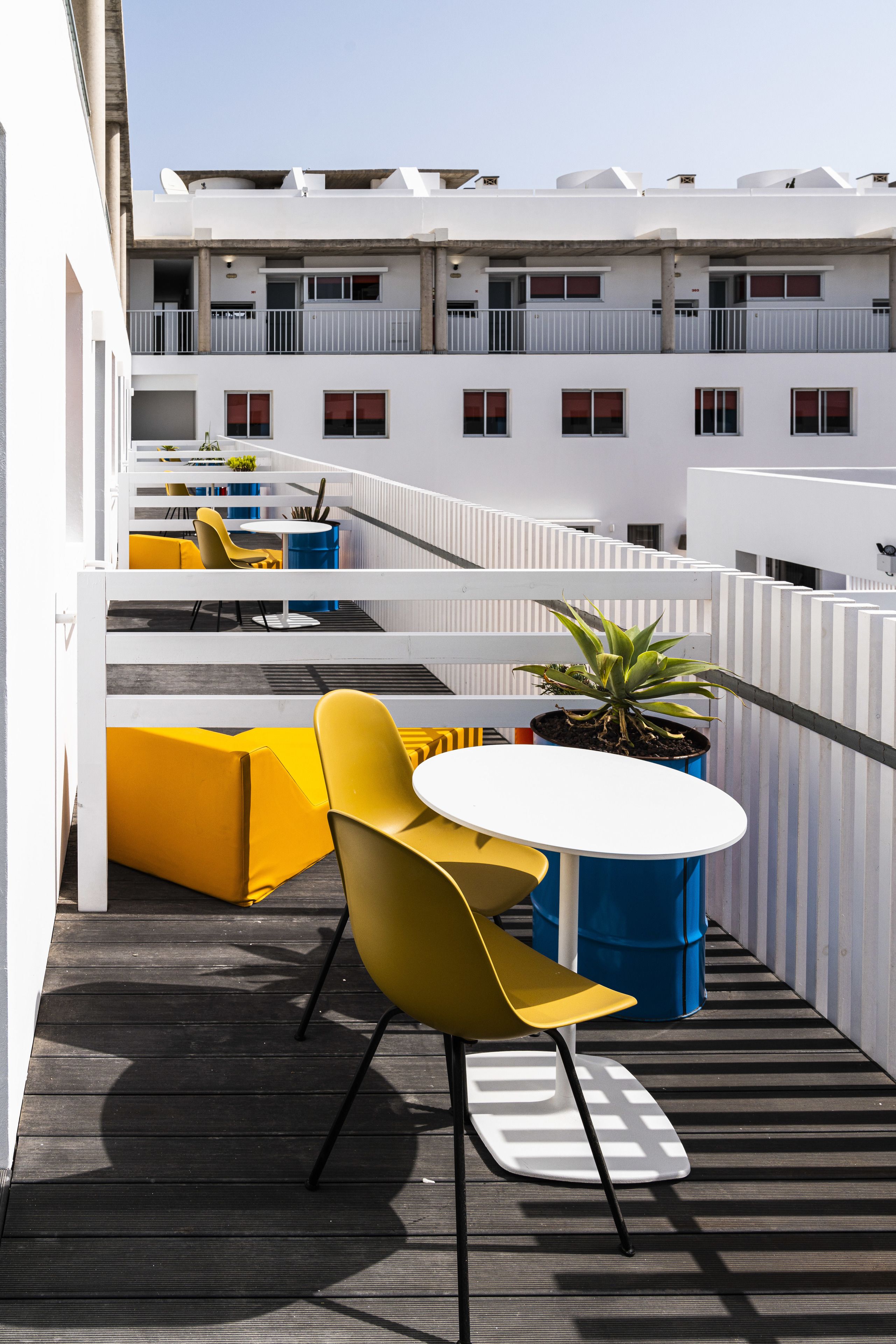 A rooftop terrace with yellow chairs and a white table, surrounded by white railings and potted plants.