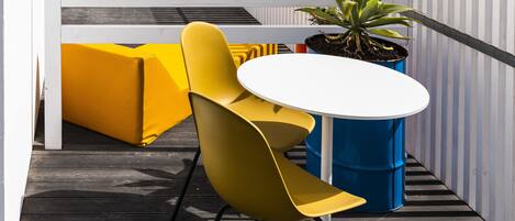 A rooftop terrace with yellow chairs and a white table, surrounded by white railings and potted plants.