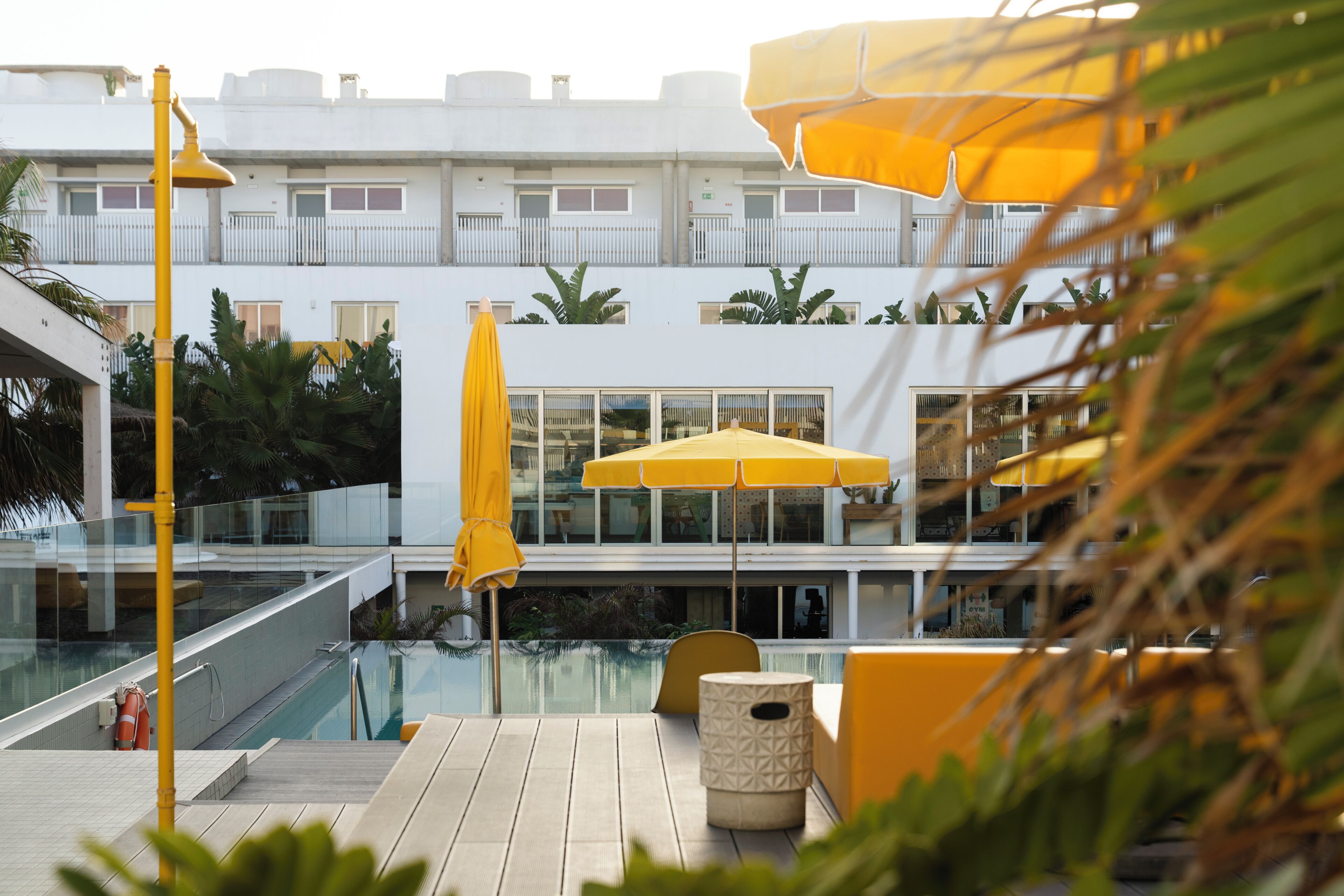 Zona de piscina con sombrillas amarillas, barandilla de cristal y un edificio blanco con balcones.