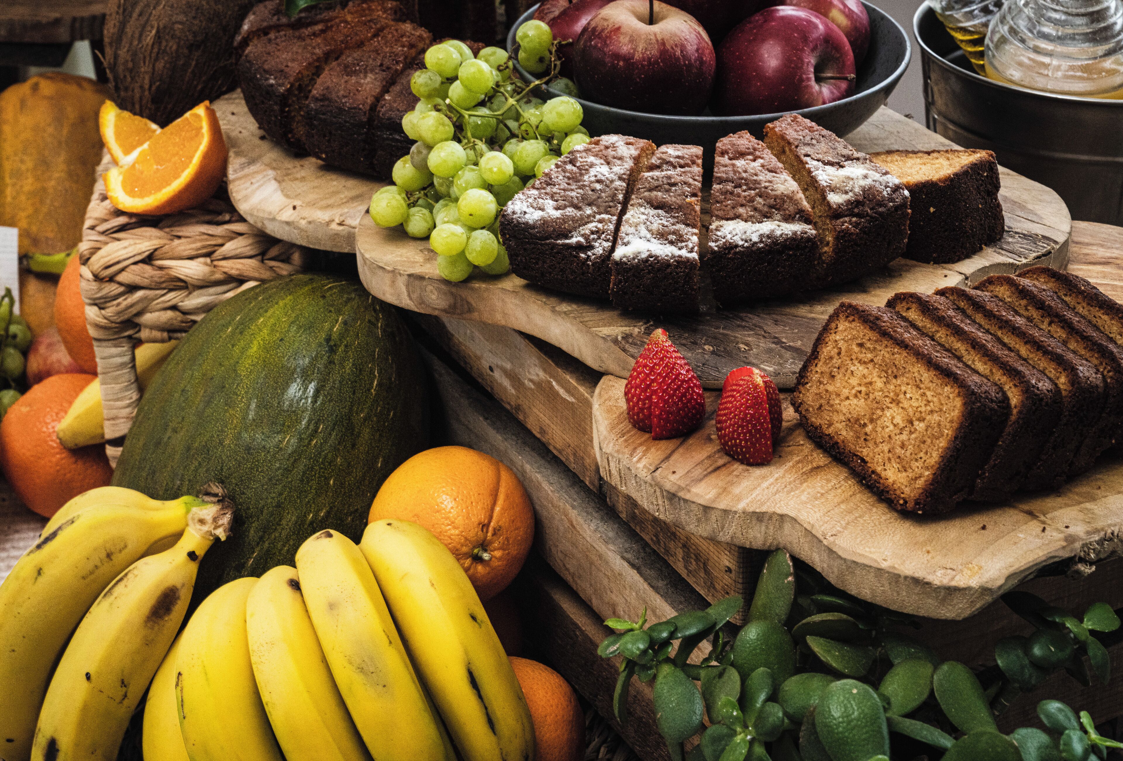 Une table en bois sur laquelle on trouve différents fruits, notamment des bananes, des oranges et des raisins, ainsi qu’une miche de pain.