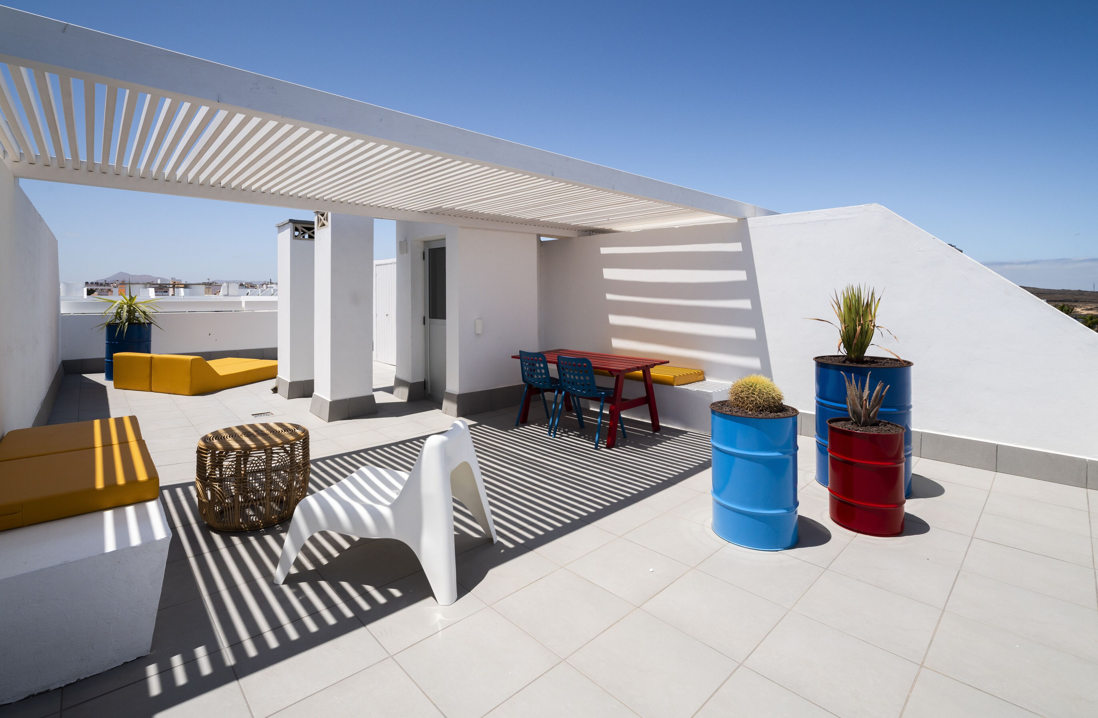 Eine Dachterrasse mit weißer Pergola, gelb-weißer Sitzgelegenheit, einem Tisch mit blauen Stühlen und Kübelpflanzen.