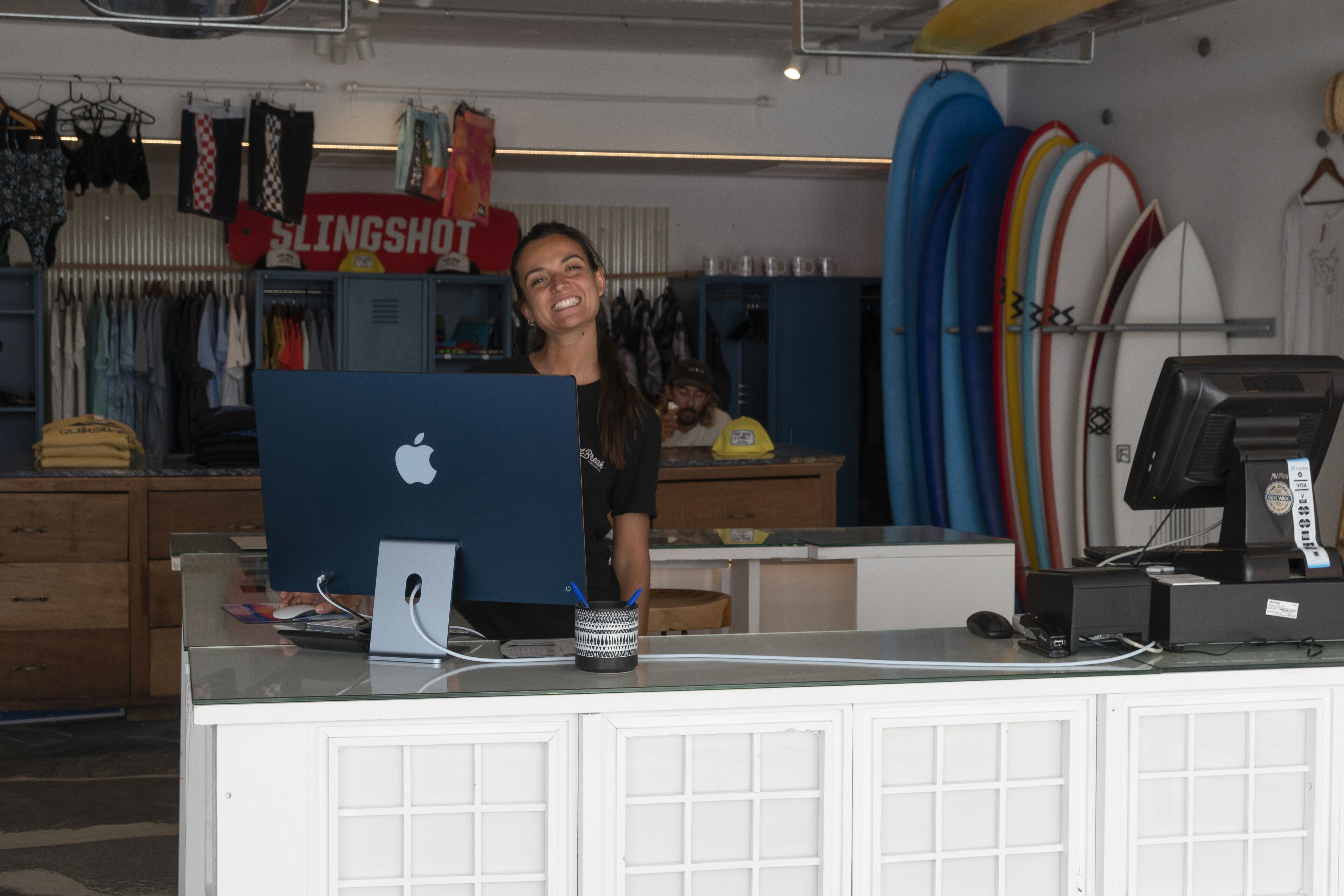Un comptoir avec un ordinateur Apple, une caisse enregistreuse et, en arrière-plan, un présentoir à planches de surf.