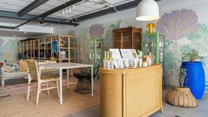 Une pièce comprenant une table à manger, des chaises, une petite table d’appoint en osier sur laquelle sont disposés des produits, un grand pot de fleurs bleu et une fresque murale.