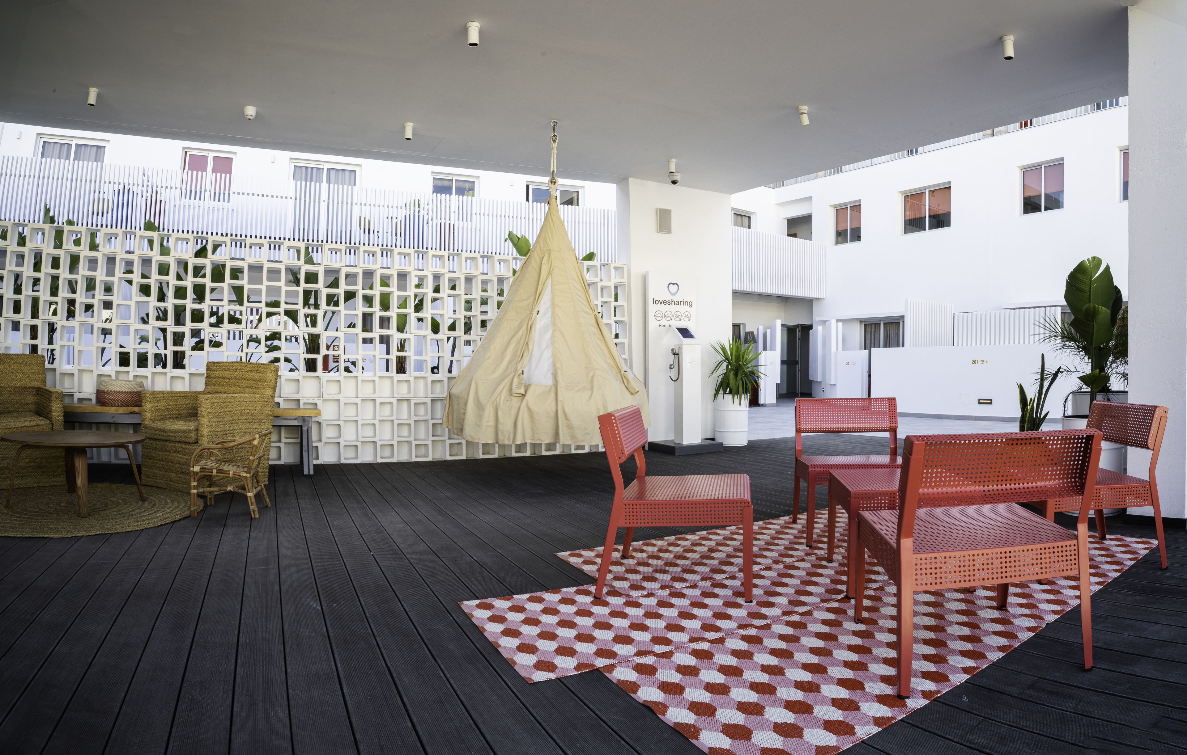Un espacio al aire libre techado con piso de madera, sillas metálicas rojas, un tapete estampado y una partición blanca con una toldo colgante.