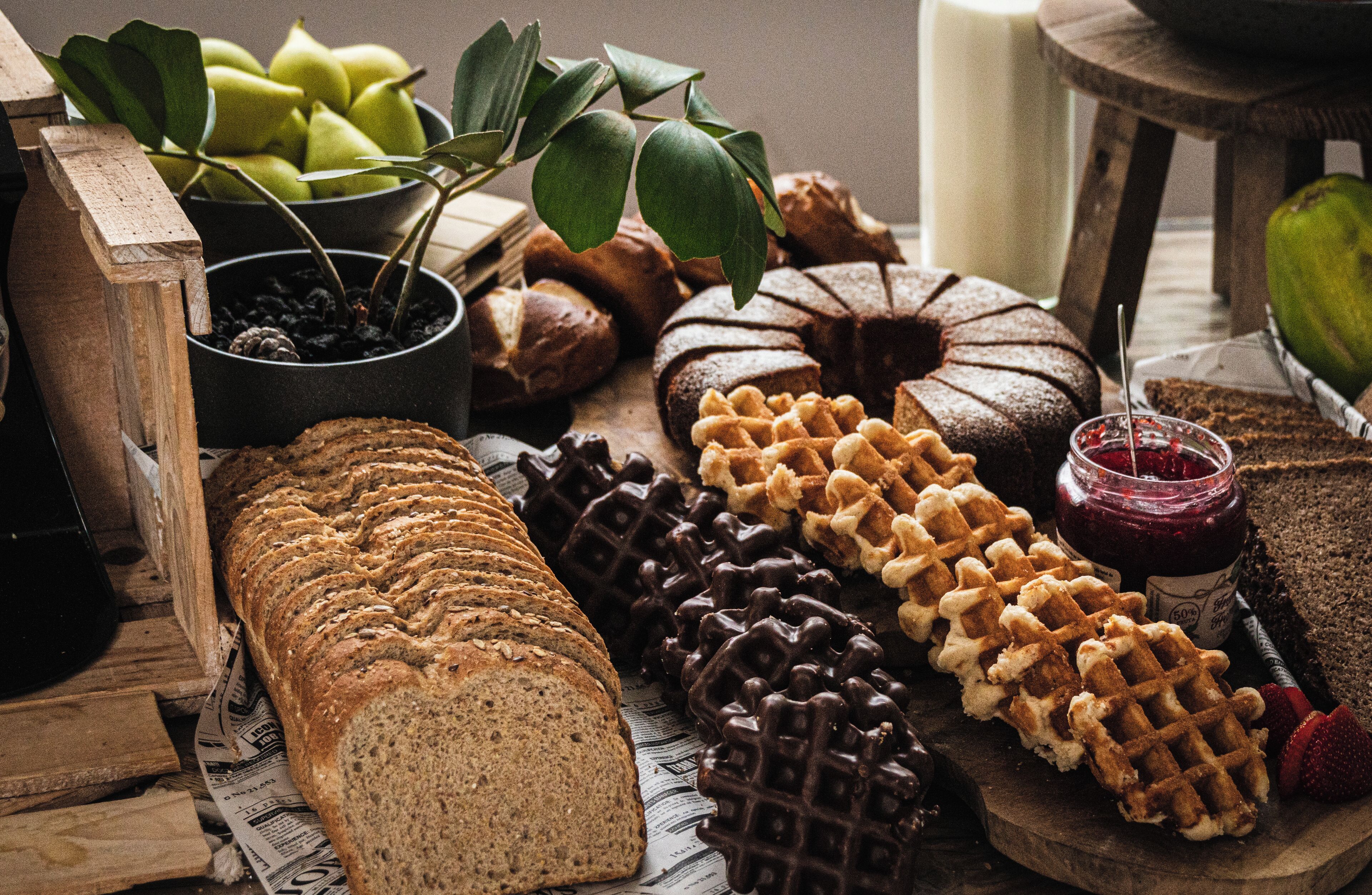 Diverse broden, wafels en gebak op een houten tafel, met een plant en een pot jam.