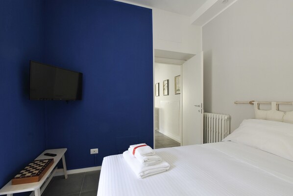 A hotel room with a bed, a bench with a chessboard, a television mounted on the wall, and a door leading to another room.