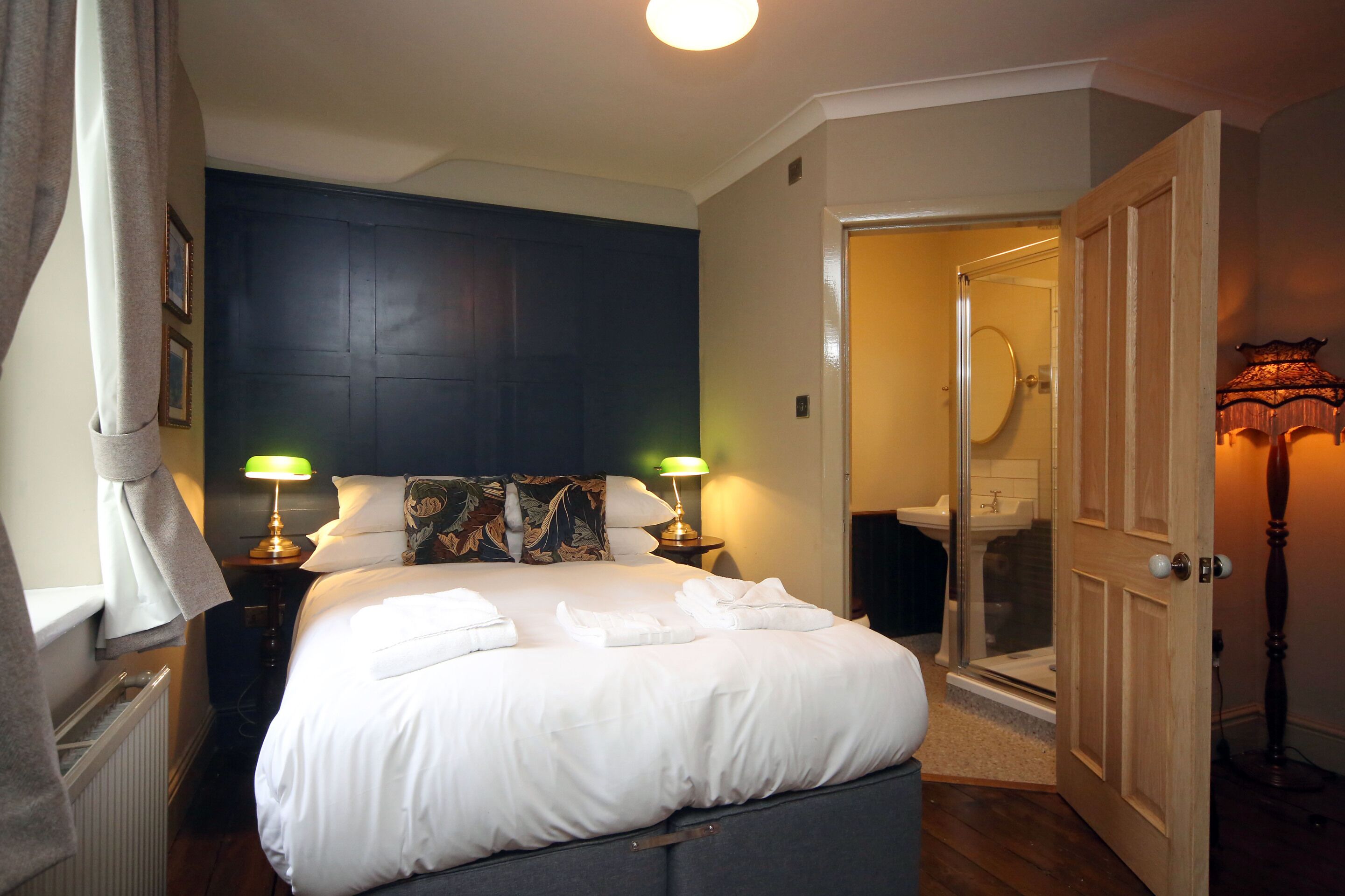 A neatly made bed with white linens and towels, a dark headboard, two bedside lamps, and a door leading to a bathroom with a glass shower enclosure.