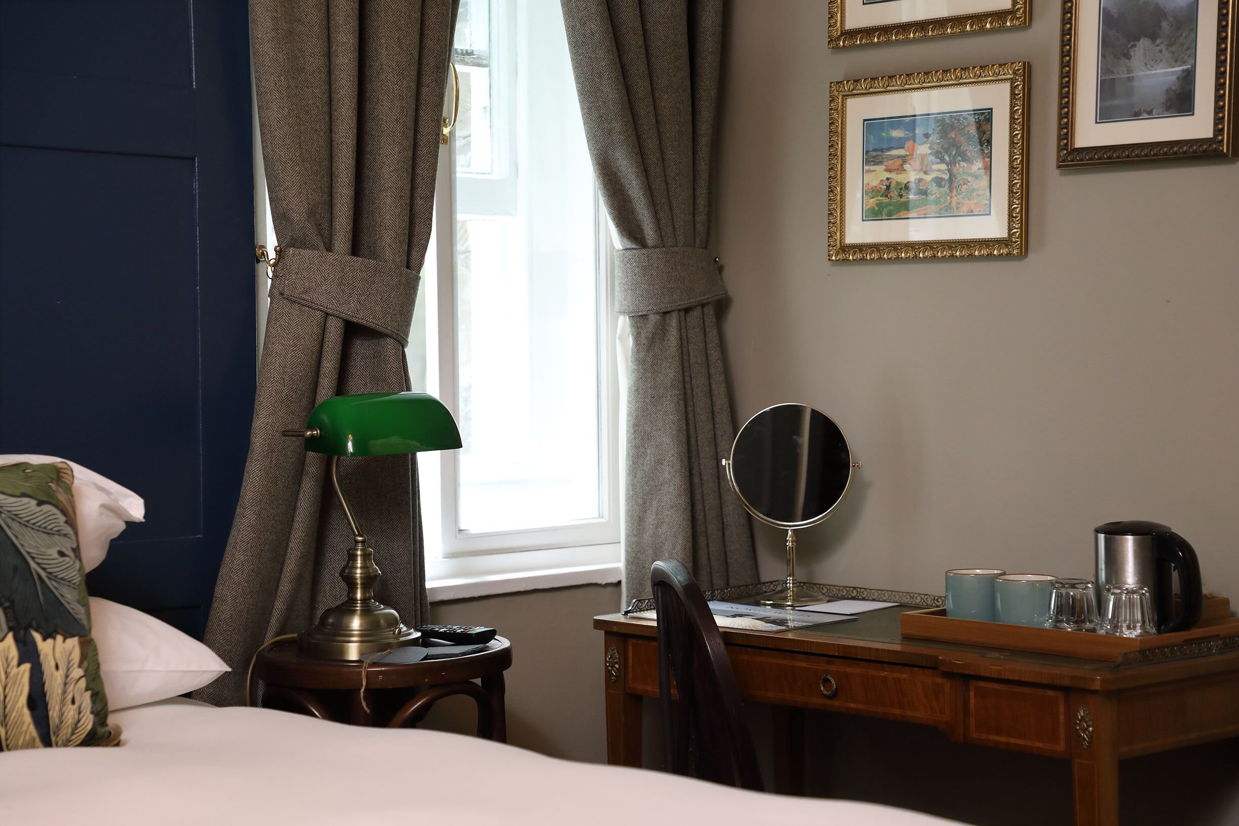 A hotel room with a bed, a desk with a lamp, a chair, a bedside table, a mirror, and framed pictures on the wall.