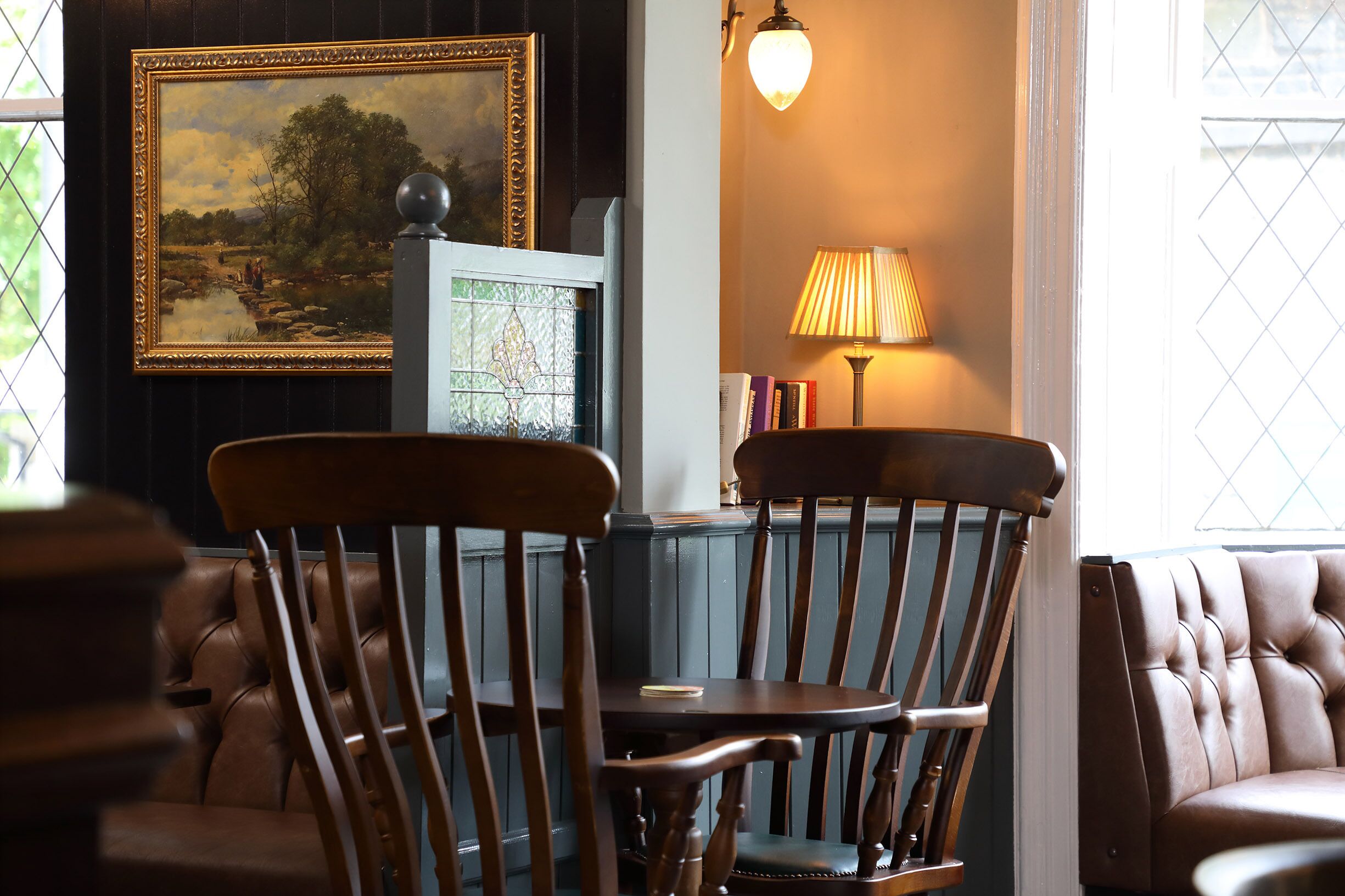 A room with a wooden chair, a table, a lamp, a framed painting, and a window with a view of greenery.
