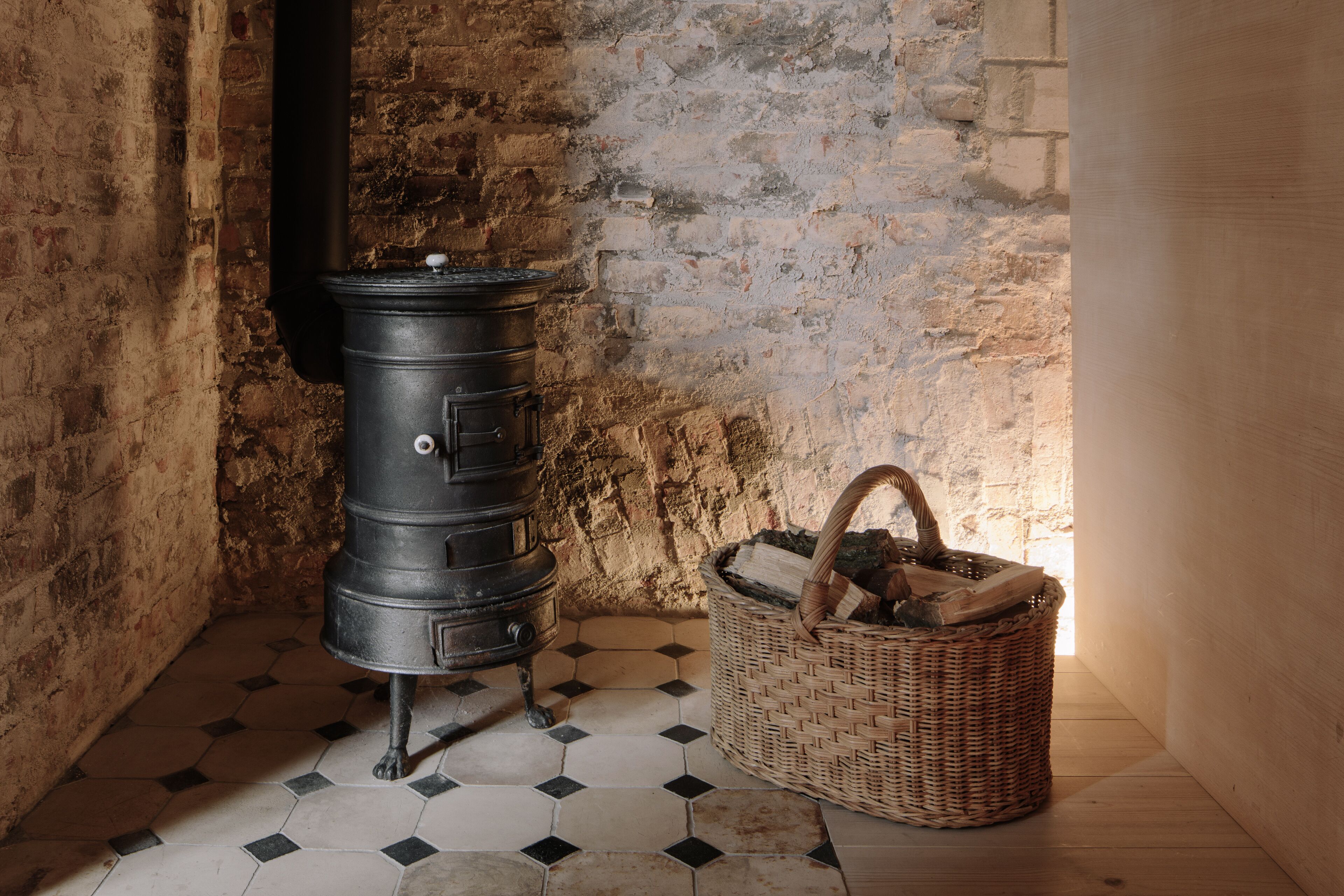 A room with a black stove, a wicker basket with firewood, and a tiled floor.