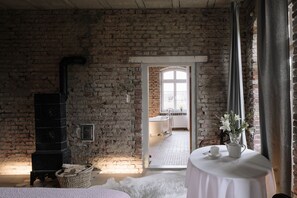 A rustic room with exposed brick walls, a fireplace, a round table with a teapot and cups, and a view into a bathroom with a bathtub and a window. - Kranich Hotel (Saal)