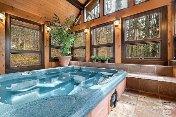 A hot tub in a wooden cabin with large windows and potted plants.
