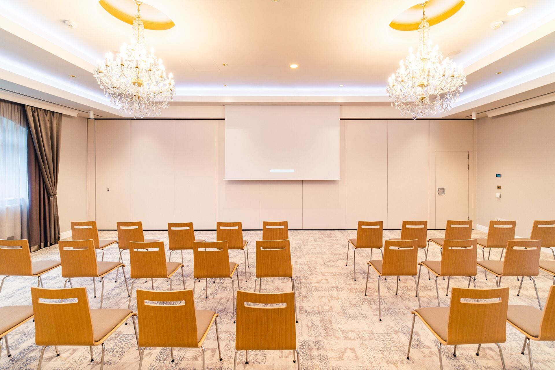 A conference room with rows of wooden chairs, a large screen, and chandeliers.