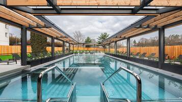 Ein Schwimmbecken mit klarem, blauem Wasser, umgeben von einer Holzterrasse und Glasgeländern, unter einer Metall-Dachkonstruktion.