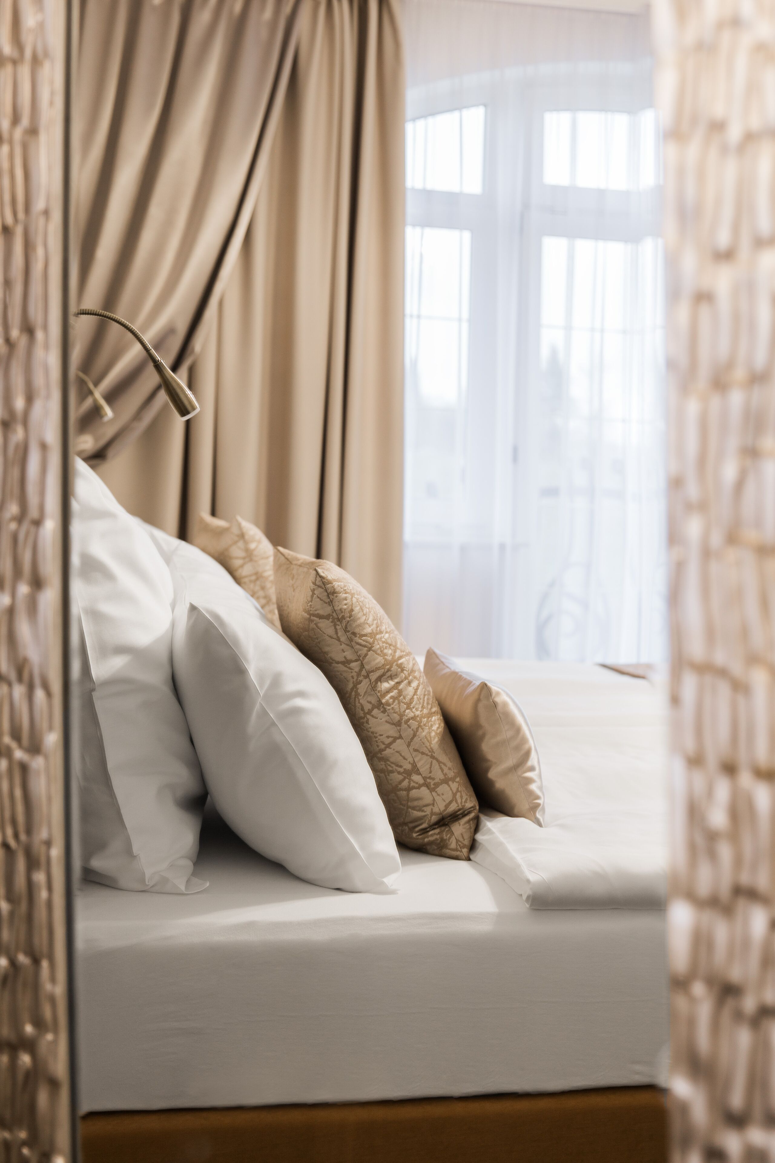 A neatly made bed with white and patterned pillows, a wooden headboard, and a view of a window with curtains.