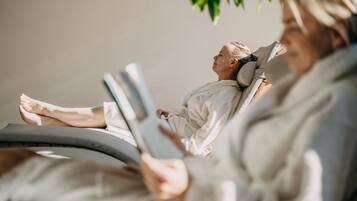Una persona recostada en una silla reclinable, leyendo un libro, con una pared lisa y una planta de fondo.