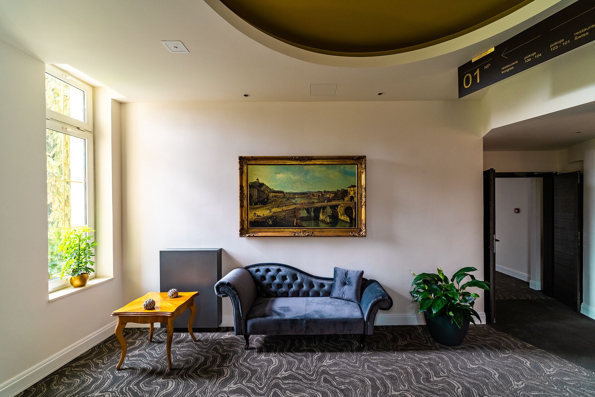 A room with a large painting of a bridge, a sofa, a small table with a plant, and a potted plant beside the sofa.