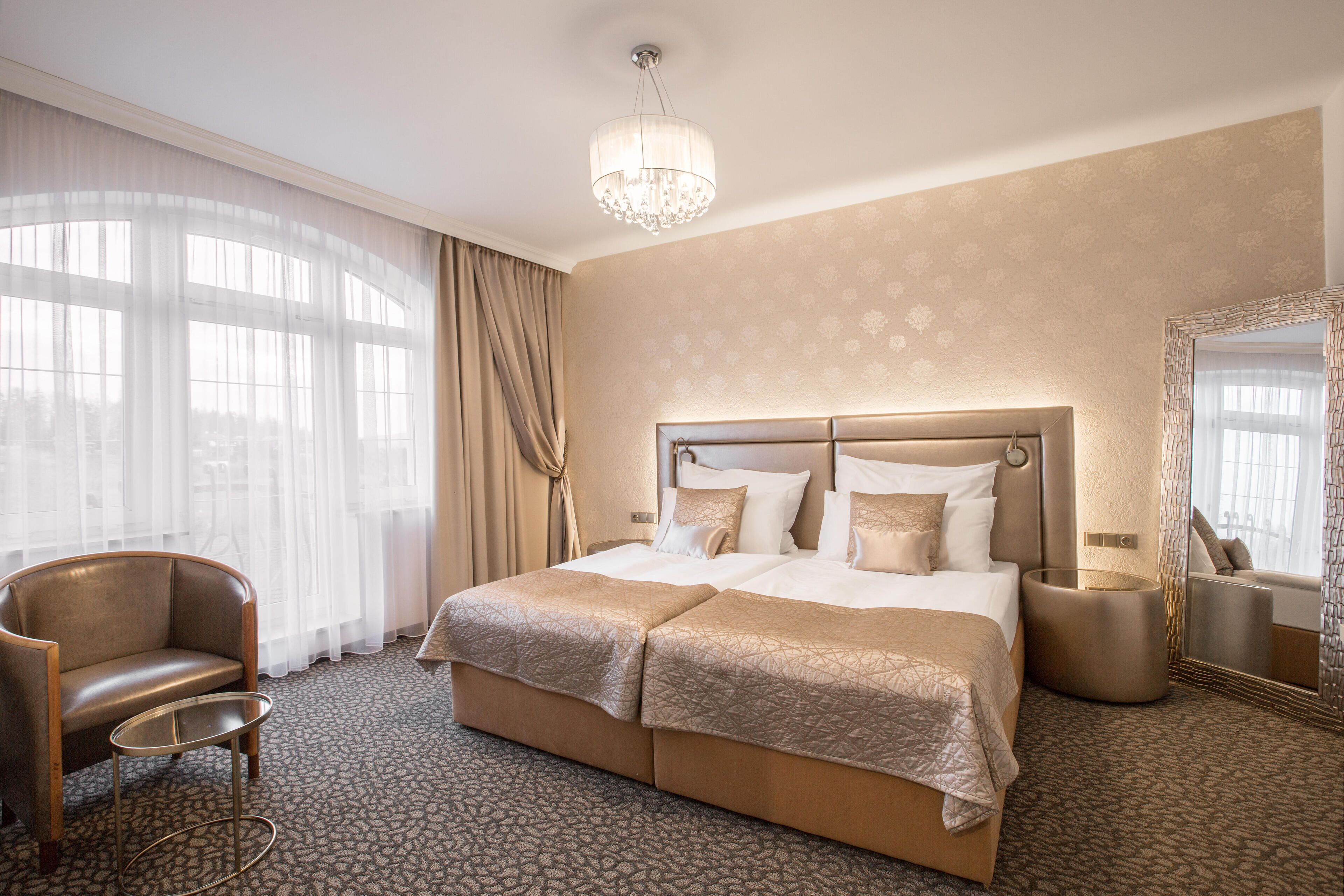 A hotel room with a large bed, a chair, a bedside table, a mirror, and a chandelier.