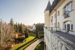 Un edificio grande e histórico con una torre destacada, un jardín cuidado con un estanque y una vista despejada del paisaje circundante.
