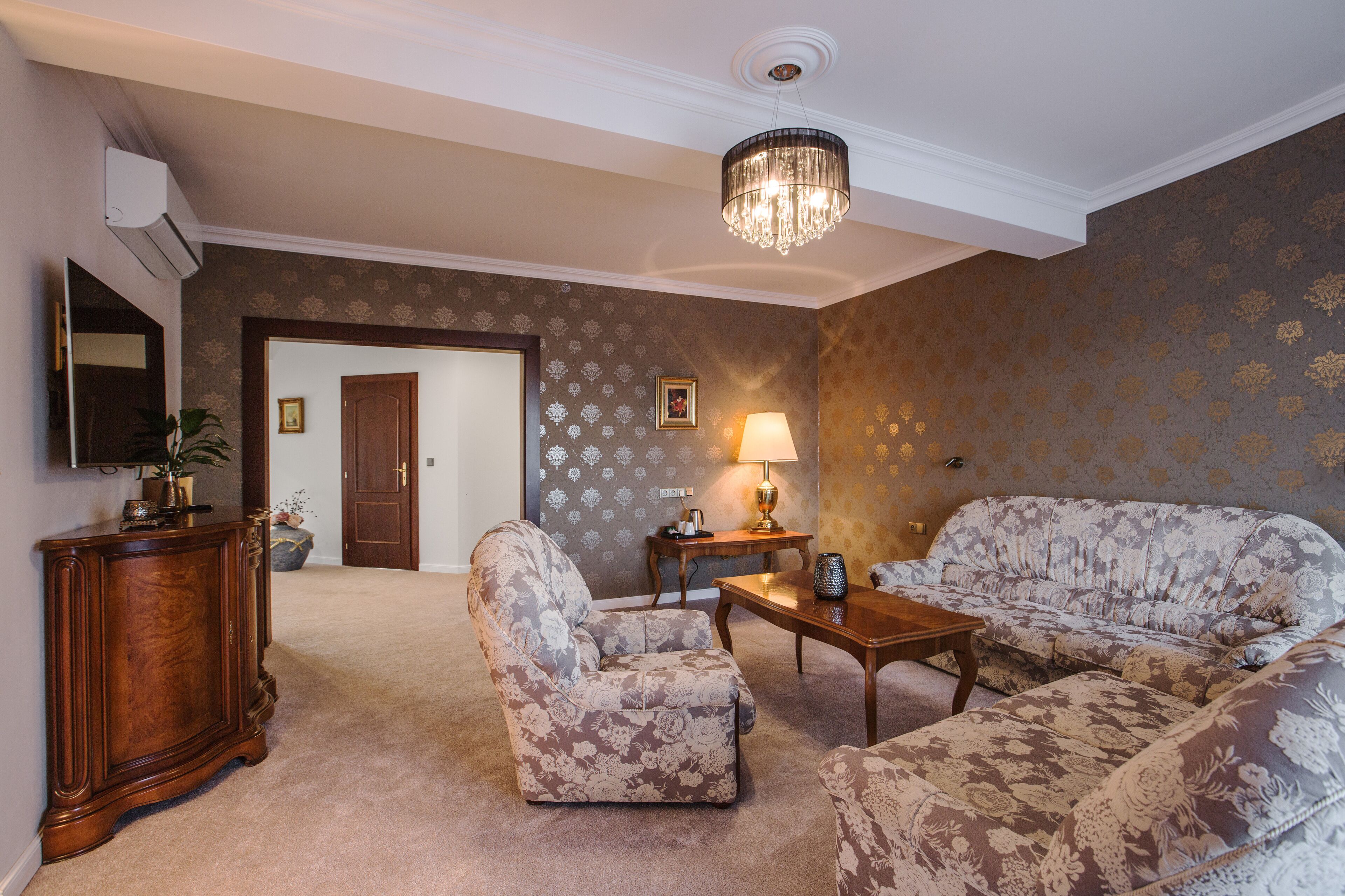 Una sala de estar con sofá, sillón y una mesa de centro de madera. Se ve una puerta que da a otra habitación, una lámpara sobre una mesita auxiliar y un jarrón decorativo.