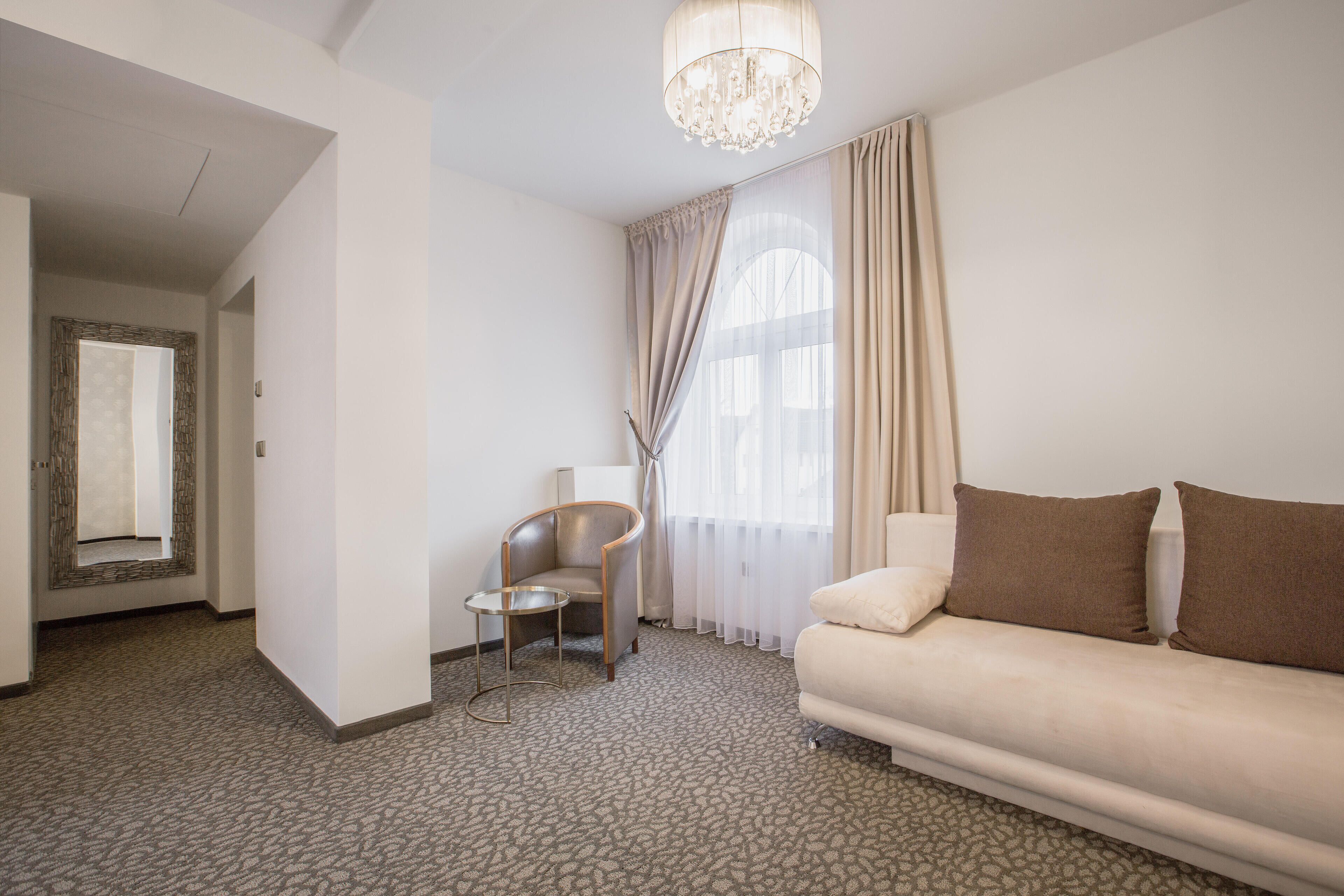 Habitación de hotel con sofá, una silla, una mesita y un ventanal con cortinas.