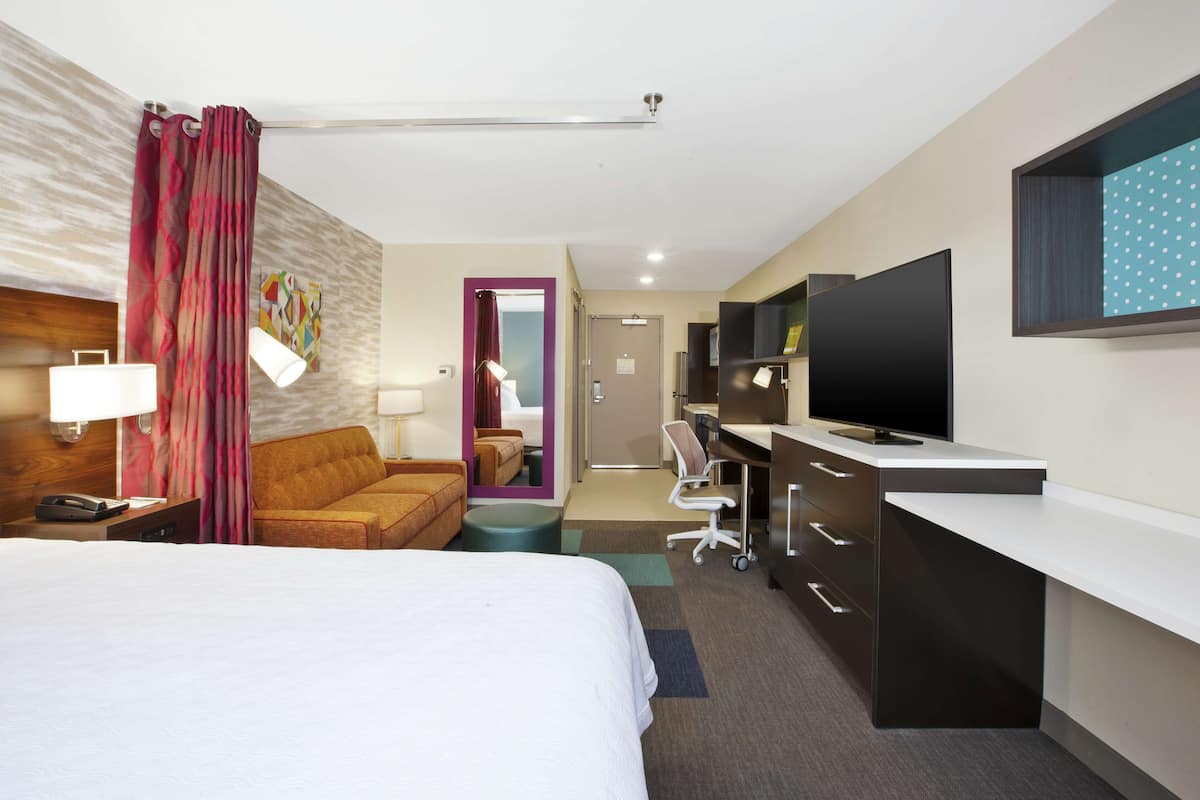 Habitación de hotel con una cama grande, un escritorio con televisor de pantalla plana, un sofá, una lámpara de escritorio y un baño visible a través de una puerta abierta.