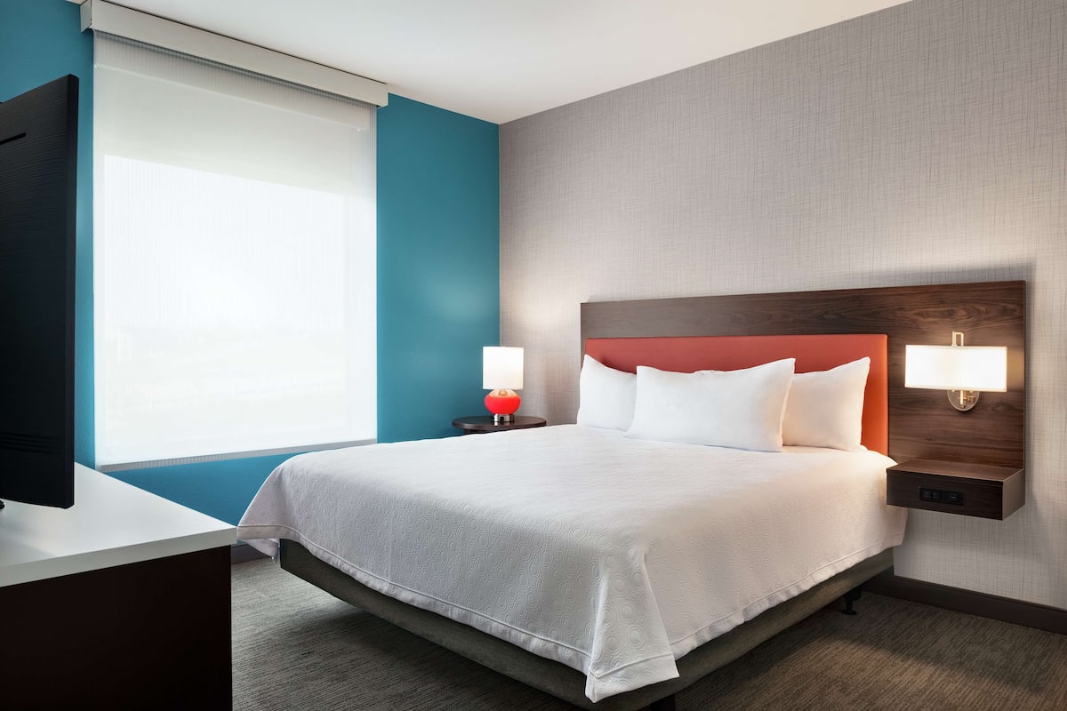 Een hotelkamer met een bed, nachtlampjes, een televisie en een raam met jaloezieën.