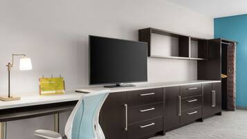 A modern hotel room with a large flat-screen TV, a desk with a lamp, a chair, and a wardrobe with multiple drawers and shelves.