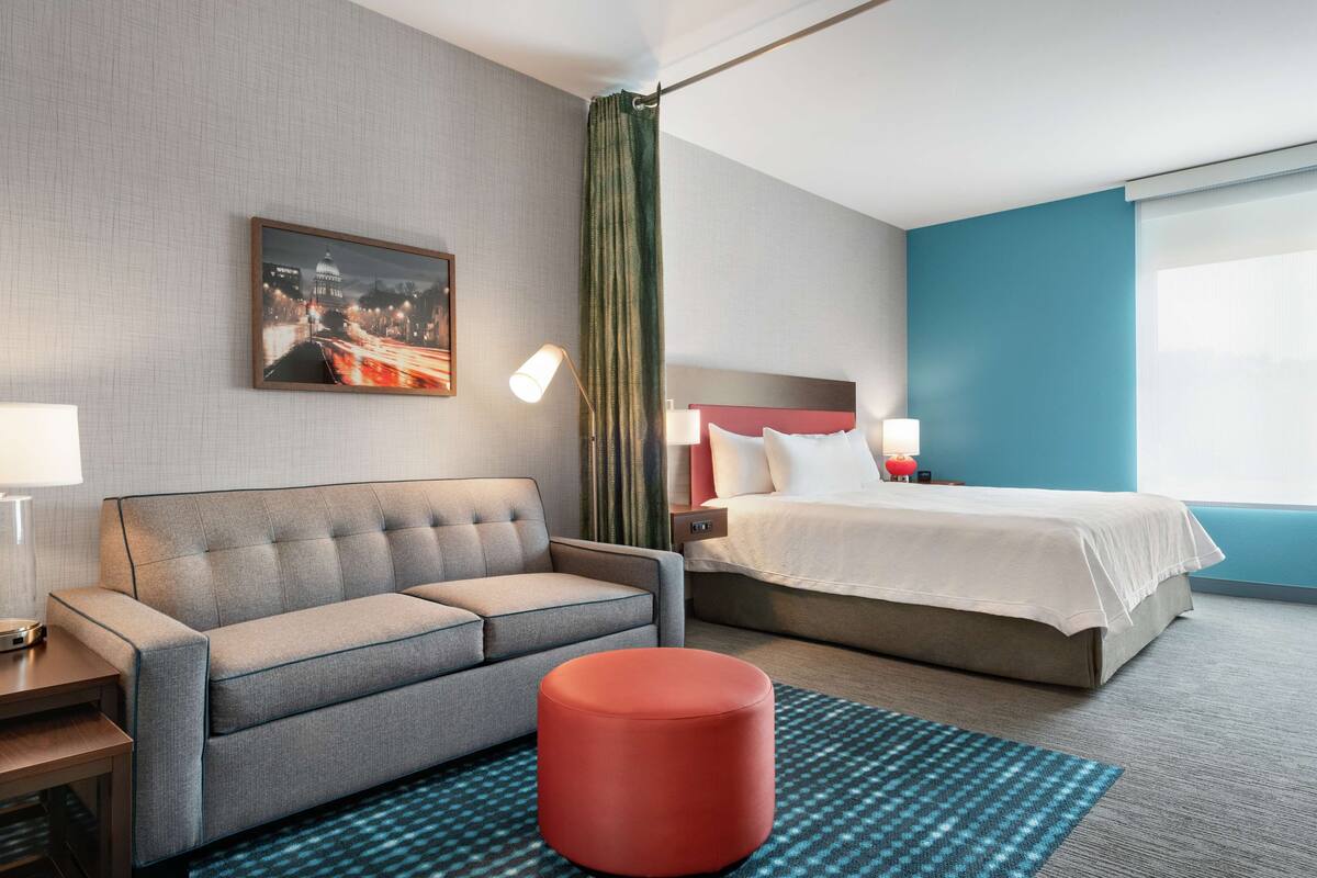 Hotelkamer met een grijze bank, een rode poef, een bed met witte lakens, een stadsgezicht schilderij aan de muur en een groot raam met gordijnen.