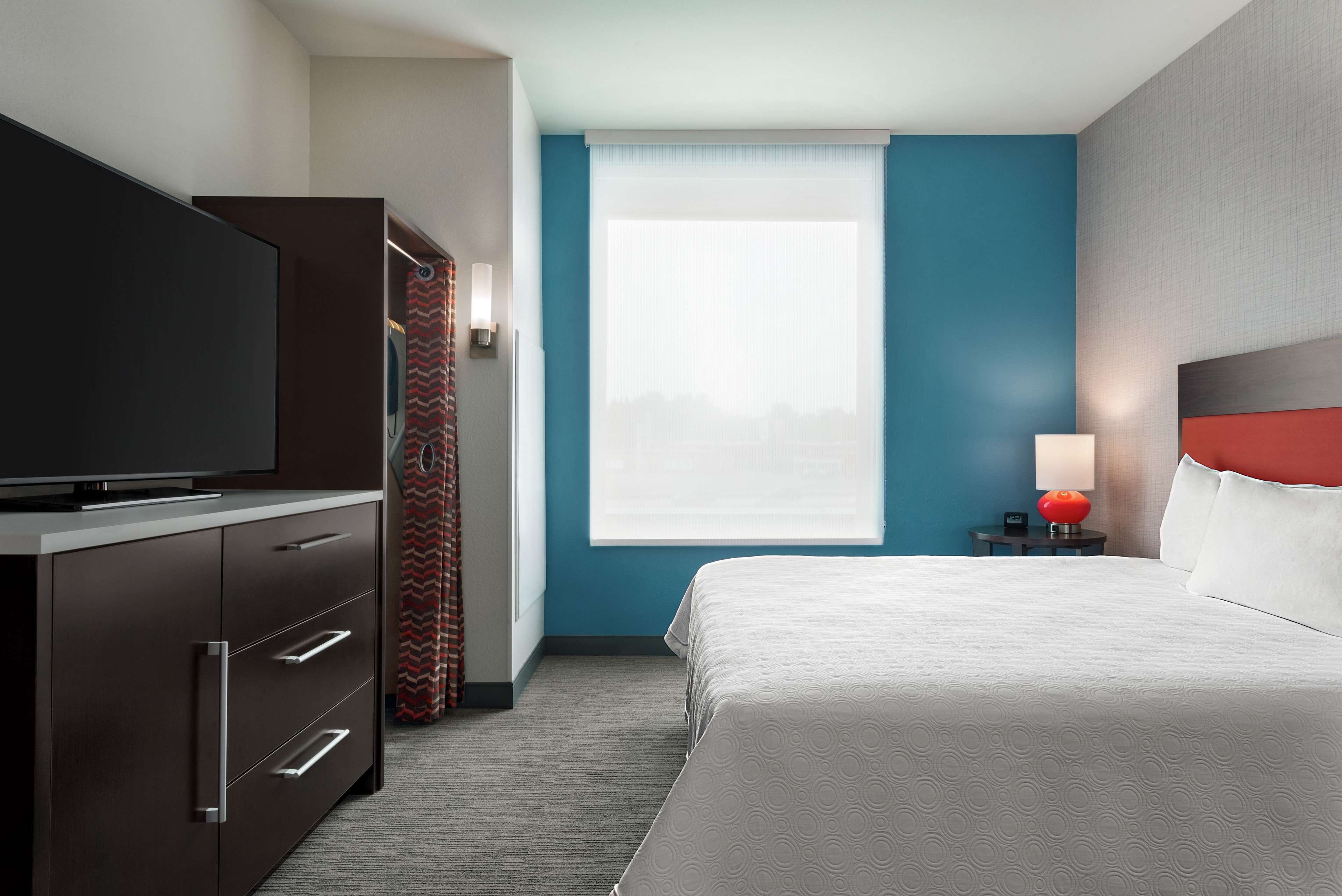 A hotel room with a bed, a TV on a cabinet, a window with curtains, and a nightstand with a lamp.