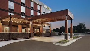 Hotellissa on katettu sisäänkäynti, ja sen yläpuolella on kyltti, jossa lukee ”HOME 2 SUITES”, sekä pysäköintialue.
