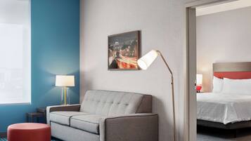 Una habitación de hotel con un sofá gris, un reposapiés rojo, un cuadro enmarcados con una vista urbana, una lámpara de noche y una cama con ropa blanca.